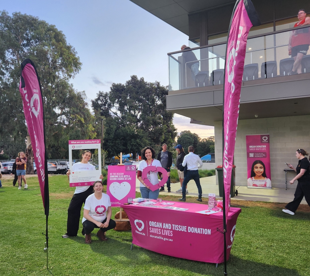 An outdoor DonateLife information stall is set up on a grassy area beside a building at dusk. Three volunteers stand behind a magenta table covered with brochures and promotional materials, wearing white DonateLife shirts. Tall magenta DonateLife flags frame the stall, and display signs include messages such as ‘Be the reason someone else gets a second chance at life’ and a photo board prop. Other people walk nearby, and an additional banner about organ donation is visible on the building in the background.