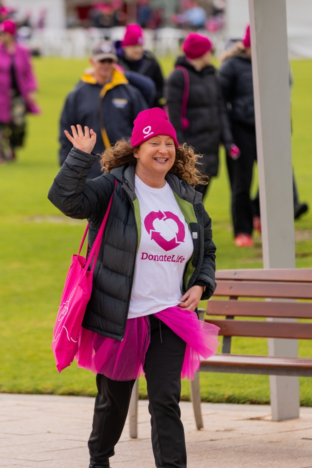 A person walks outdoors wearing a pink beanie, white 'DonateLife' shirt, black jacket and pants, and a pink tutu, waving and carrying a pink bag. Others in the background are dressed warmly, some also wearing pink beanies. 