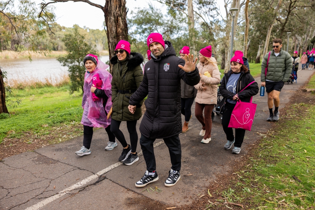 A group of people walk along a paved path in a park, dressed in warm clothing including bright pink beanies, ponchos, and bags. Trees and greenery surround the path, with a body of water visible on the left.