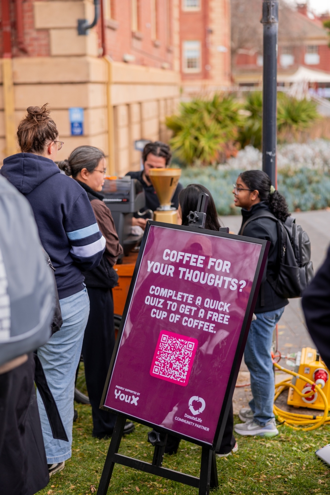 A group of people stand near a coffee stand with a sign in the foreground that reads 'COFFEE FOR YOUR THOUGHTS? Complete a quick quiz to get a free cup of coffee.' The sign includes a QR code and logos for 'YouX' and 'DonateLife Community Partner.'