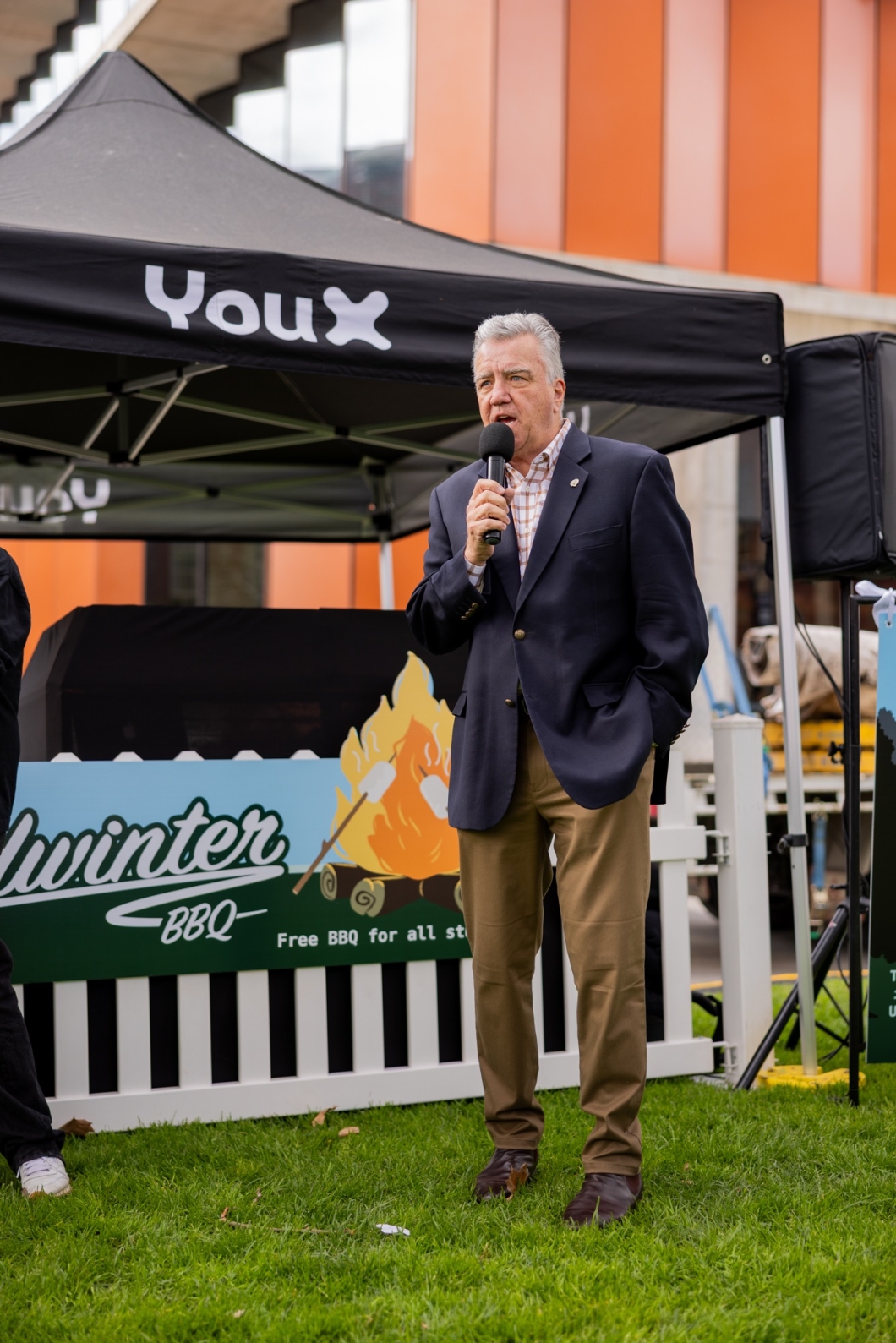 A man stands on grass holding a microphone and speaking, dressed in a navy blazer, light shirt, and khaki pants. Behind him is a black canopy tent labeled 'YouX' and a sign that reads 'Winter BBQ' with an illustration of marshmallows roasting over a fire and the text 'Free BBQ for all students’.