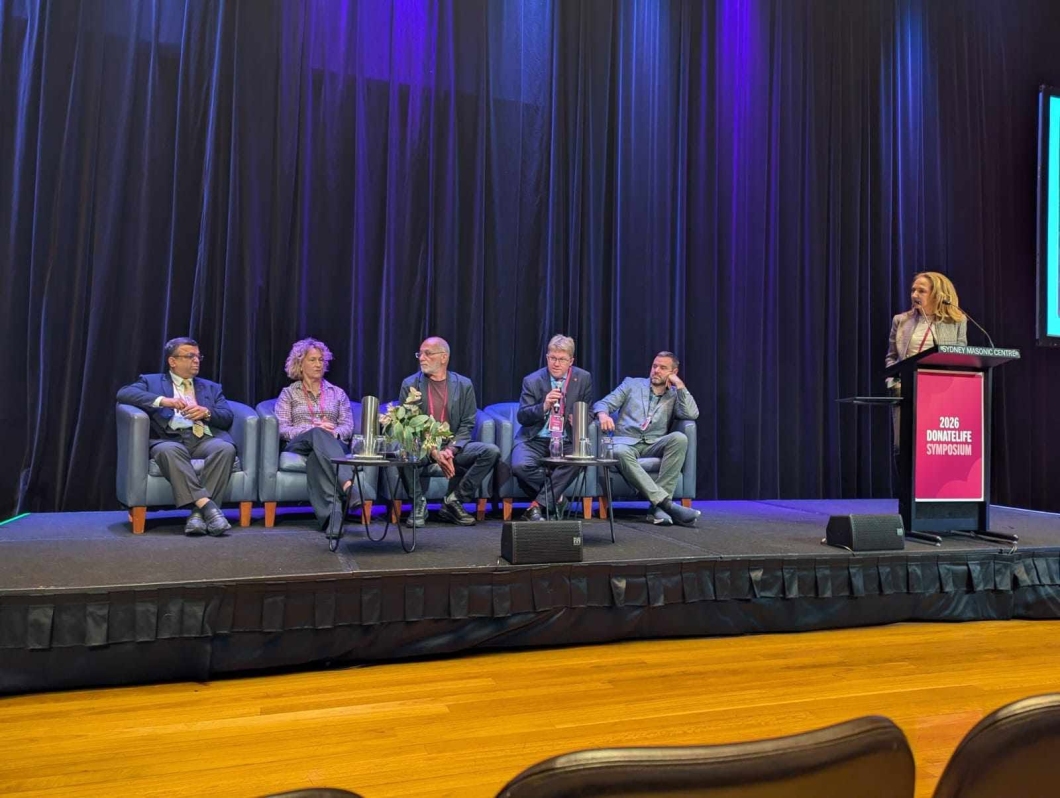 A panel of speakers sit on stage during the 2026 DonateLife Symposium, while a moderator stands at a lectern leading a discussion in front of an audience.