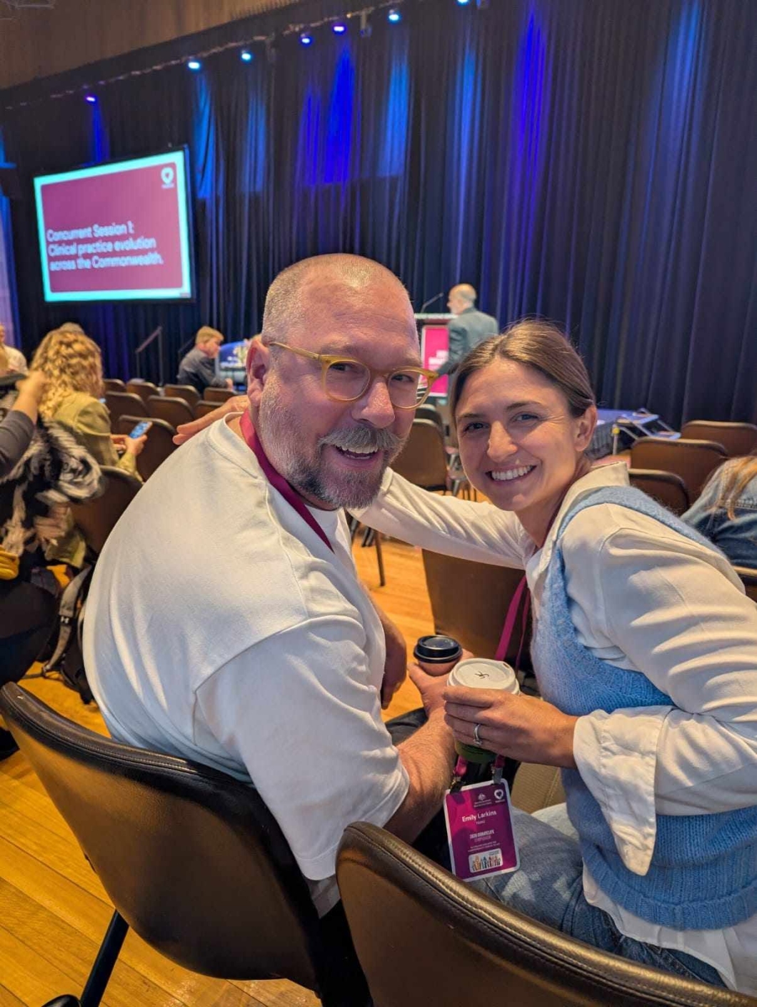 2 symposium delegates sit together in an auditorium before a presentation, wearing event lanyards and holding takeaway coffee cups.