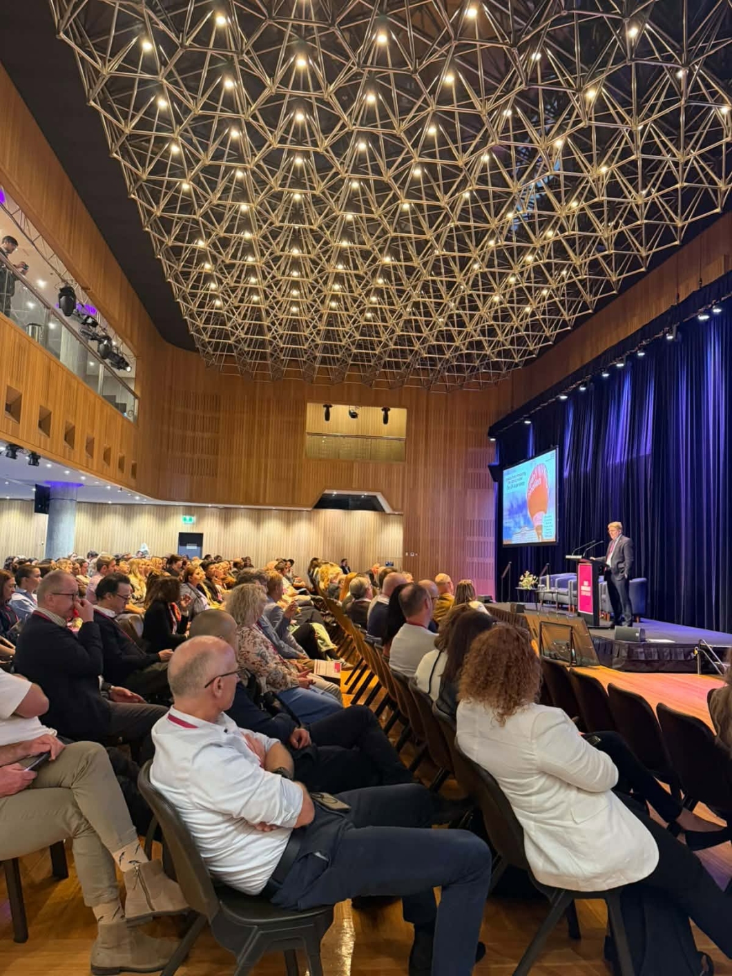 Delegates seated in a large auditorium listen to a speaker presenting on stage during the 2026 DonateLife Symposium, beneath an illuminated geometric ceiling feature.