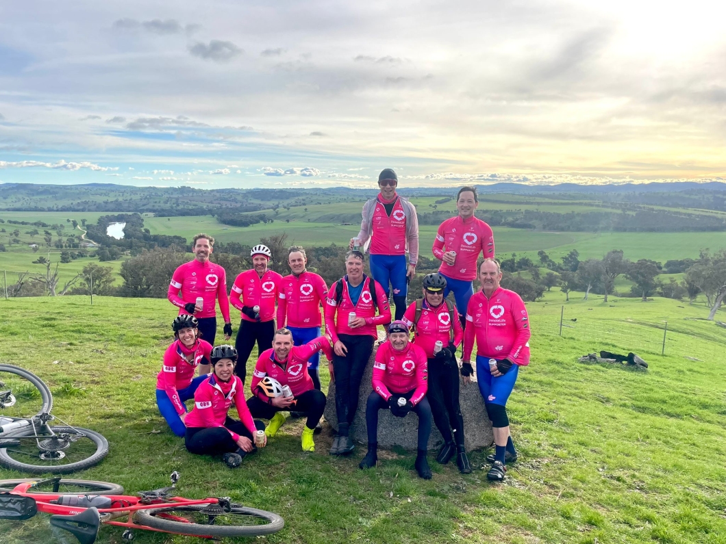 12 cyclists in matching magenta jerseys pose on a grassy hilltop with bicycles nearby, overlooking green fields, trees, and distant hills under a partly cloudy sky.