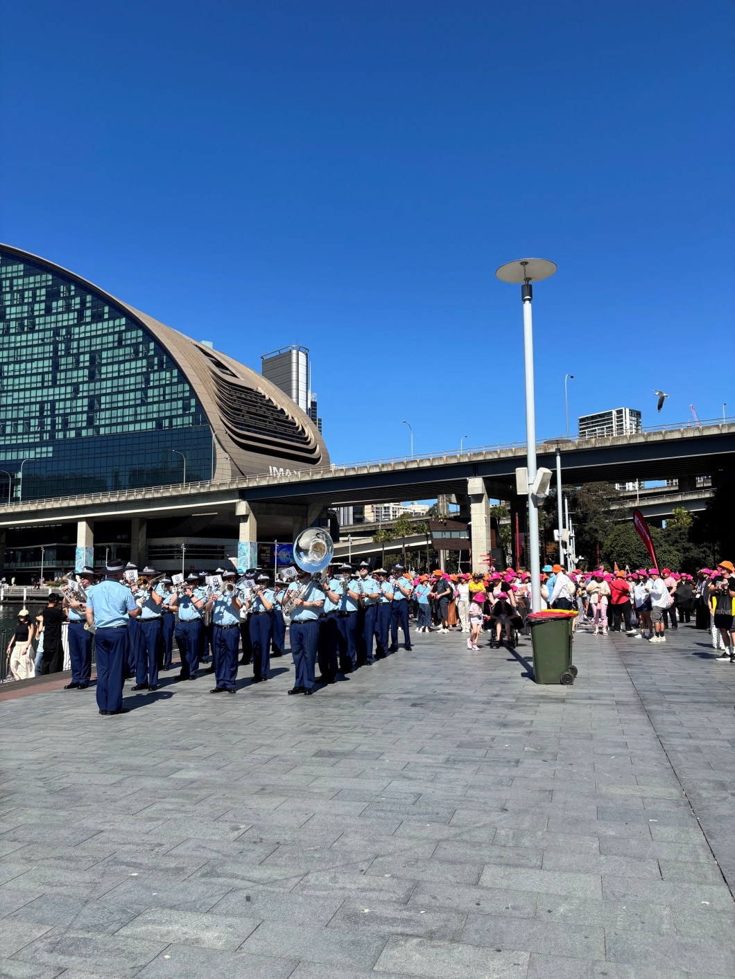 A marching band leads participants wearing bright hats through a waterfront precinct during a community walk raising awareness of organ and tissue donation.