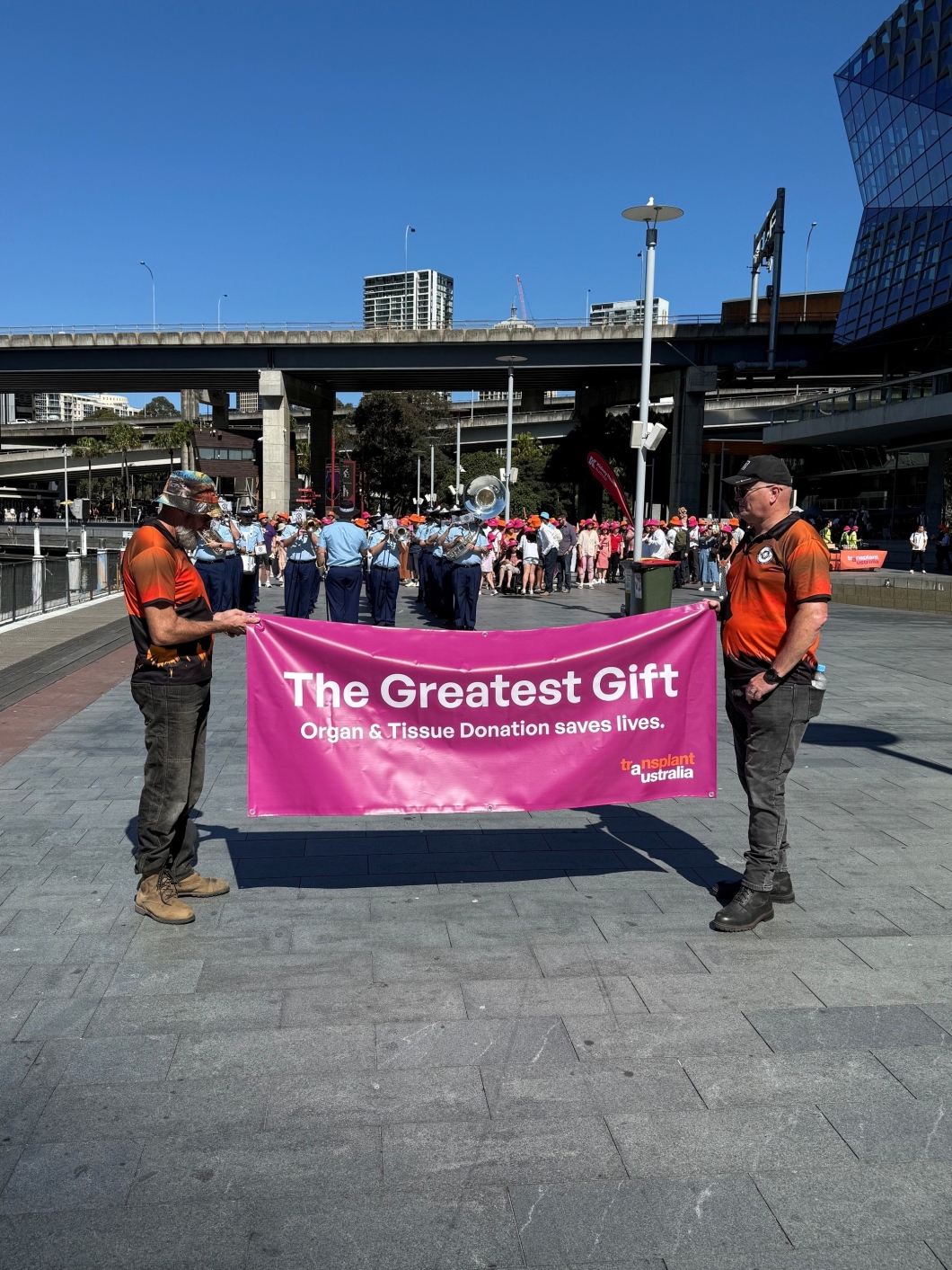 2 participants hold a large magenta banner reading ‘The Greatest Gift’ at the front of a community walk promoting organ and tissue donation, with a marching band and participants following behind.