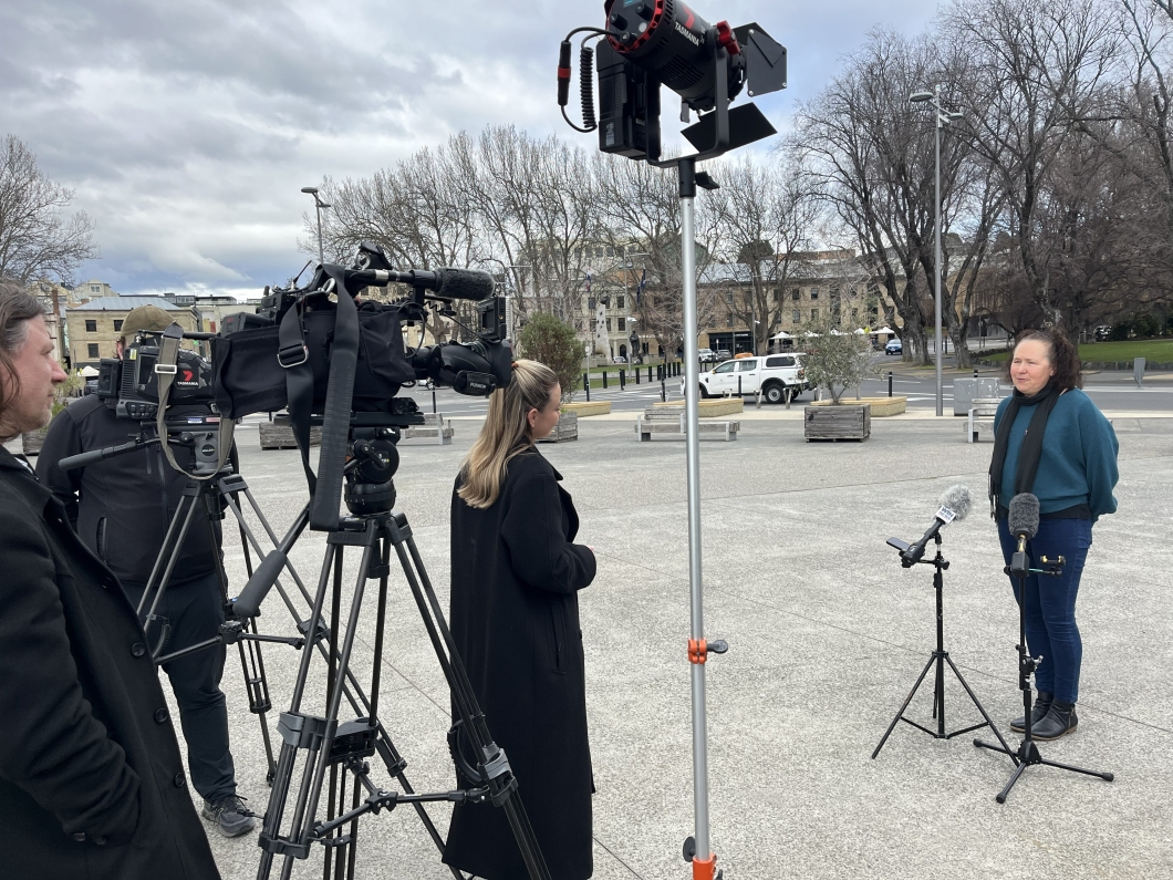 A television news crew records an outdoor interview in a public square, with a spokesperson speaking beside microphones while cameras and lighting equipment capture the media appearance during DonateLife Week.