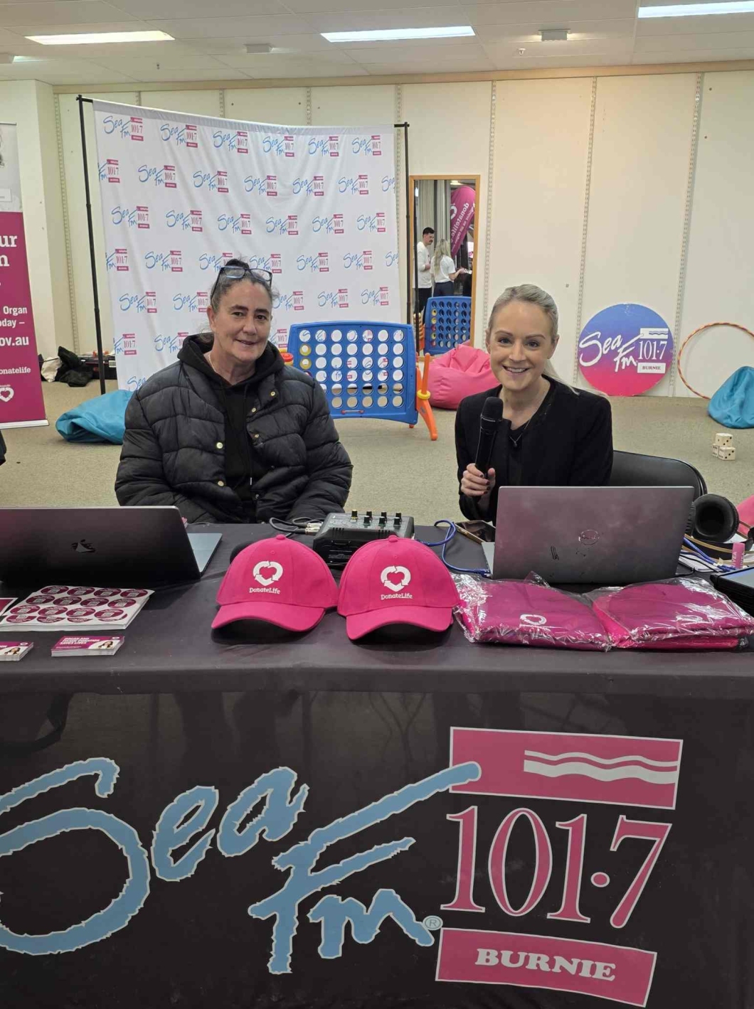 2 people sit behind a Sea FM 101.7 Burnie outside broadcast desk displaying DonateLife information, with magenta DonateLife caps and promotional materials arranged on the table.