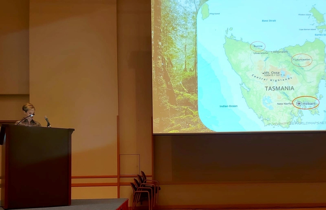 A person stands at a lectern in a conference room, presenting beside a large projected slide. The slide shows a map of Tasmania with cities including Burnie, Launceston, and Hobart circled, alongside an image of a forest on the left side of the slide.