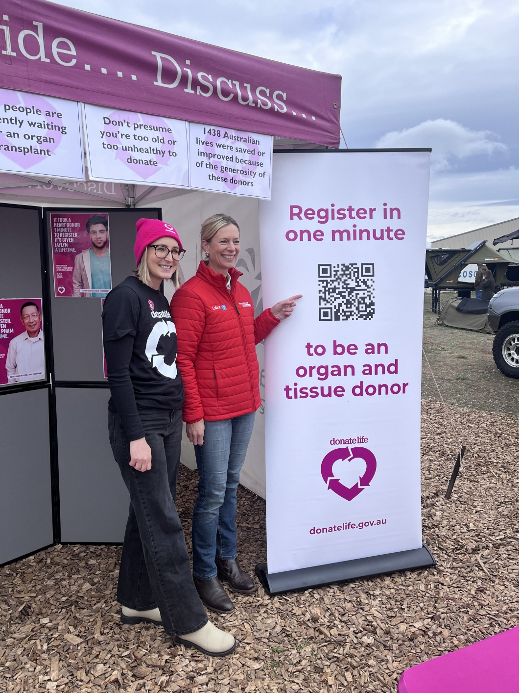 2 people stand beside a DonateLife information booth at an outdoor event, next to a tall sign that reads ‘Register in one minute to be an organ and tissue donor’ with a QR code. One person points to the QR code while informational posters about organ donation are displayed on the booth behind them under a magenta canopy.