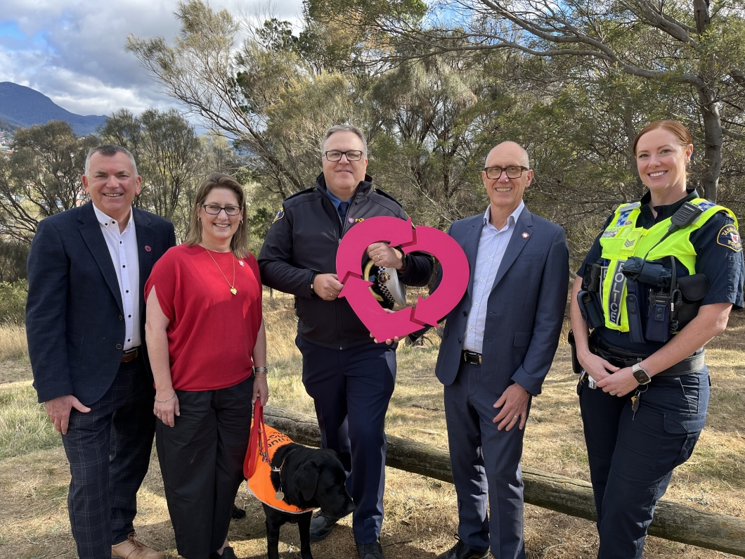 5 people stand outdoors in a bushland setting with trees and hills in the background, posing together for a group photo. One person in the centre holds a large DonateLife logo cut-out. A police officer in a high-visibility vest stands on the far right. A black dog wearing an orange vest is partially visible at the front. The group is dressed in a mix of business attire and uniforms, and several are wearing magenta pins on their clothing.