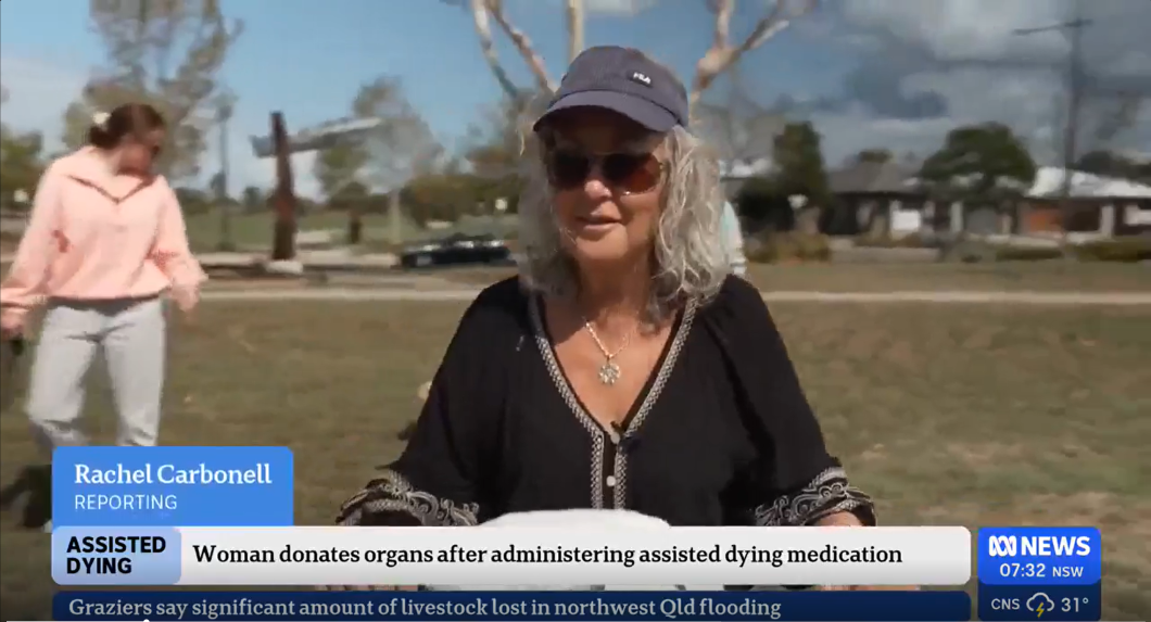 A news broadcast shows a person outdoors in a park, wearing a cap and dark top, standing in front of trees and houses. A lower-third graphic reads ‘Rachel Carbonell — Reporting’ and ‘ASSISTED DYING — Woman donates organs after administering assisted dying medication.’ The ABC News logo and the time appear in the corner.