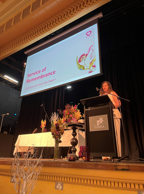 A person stands at a podium on a stage delivering a speech at a Service of Remembrance event. A large screen above displays the title ‘Service of Remembrance, Collingwood Town Hall Naarm’ with floral artwork. The stage is decorated with floral arrangements, candles, and a white-draped table.