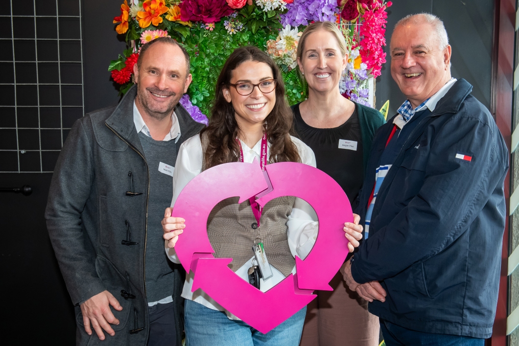 4 people stand in front of a colourful floral display, holding a large magenta DonateLife symbol during a DonateLife Week community event.