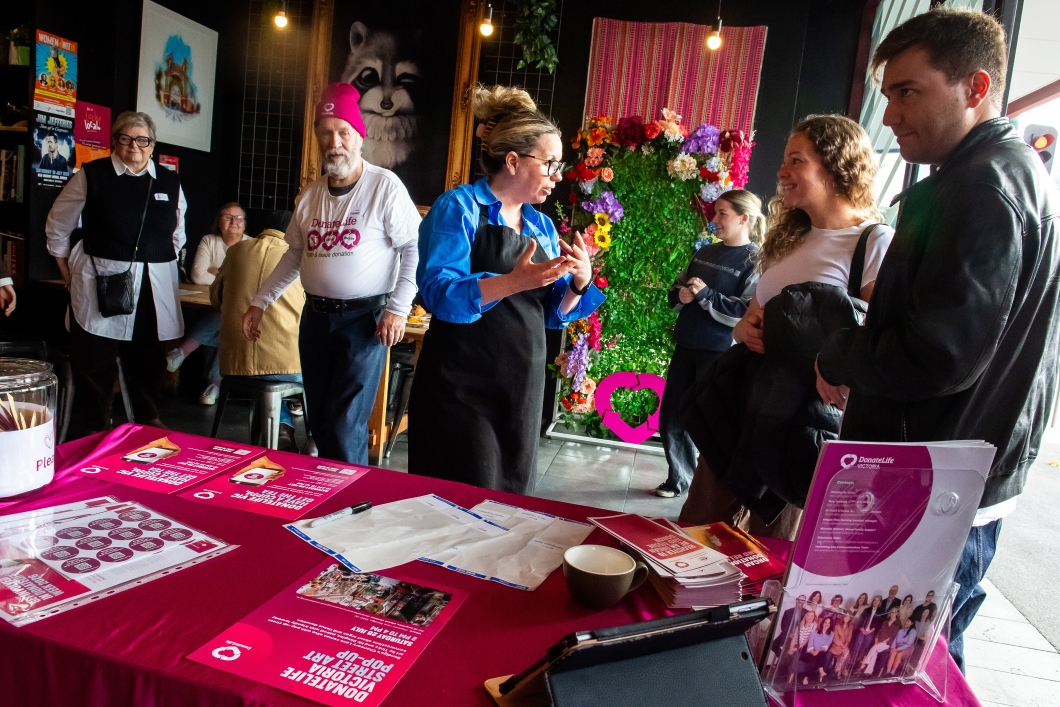 Visitors speak with DonateLife representatives at an information stall displaying magenta table coverings, brochures and registration materials during a DonateLife Week community event.