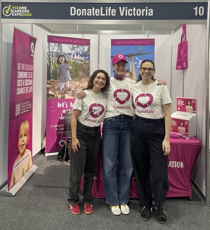 3 people stand side by side at a DonateLife Victoria exhibition booth at the VCE and Careers Expo. All wear white DonateLife shirts with a heart logo and have arms around each other. Behind them are banners and signs reading ‘DonateLife Victoria’ and ‘donatelife.gov.au,’ featuring photos of children and messages about organ donation. A magenta table and tote bags are visible inside the booth.