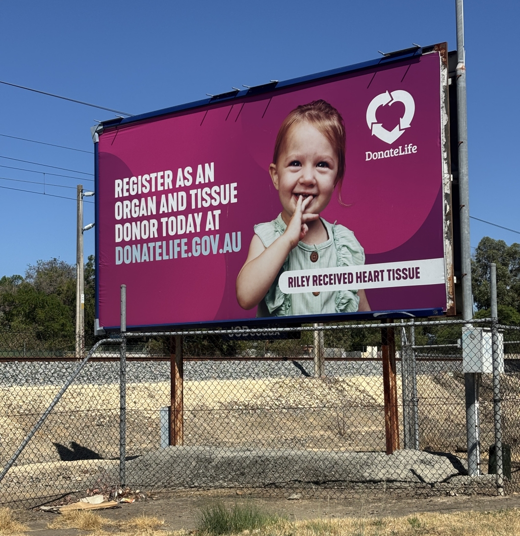 A large roadside billboard displays a DonateLife campaign featuring a young child holding their hand to their chest. The billboard has a bright pink background with the text ‘Register as an organ and tissue donor today at donatelife.gov.au’ and a label reading ‘Riley received heart tissue.’ The DonateLife logo appears in the top right. A wire fence and open, dry ground sit in front of the billboard.
