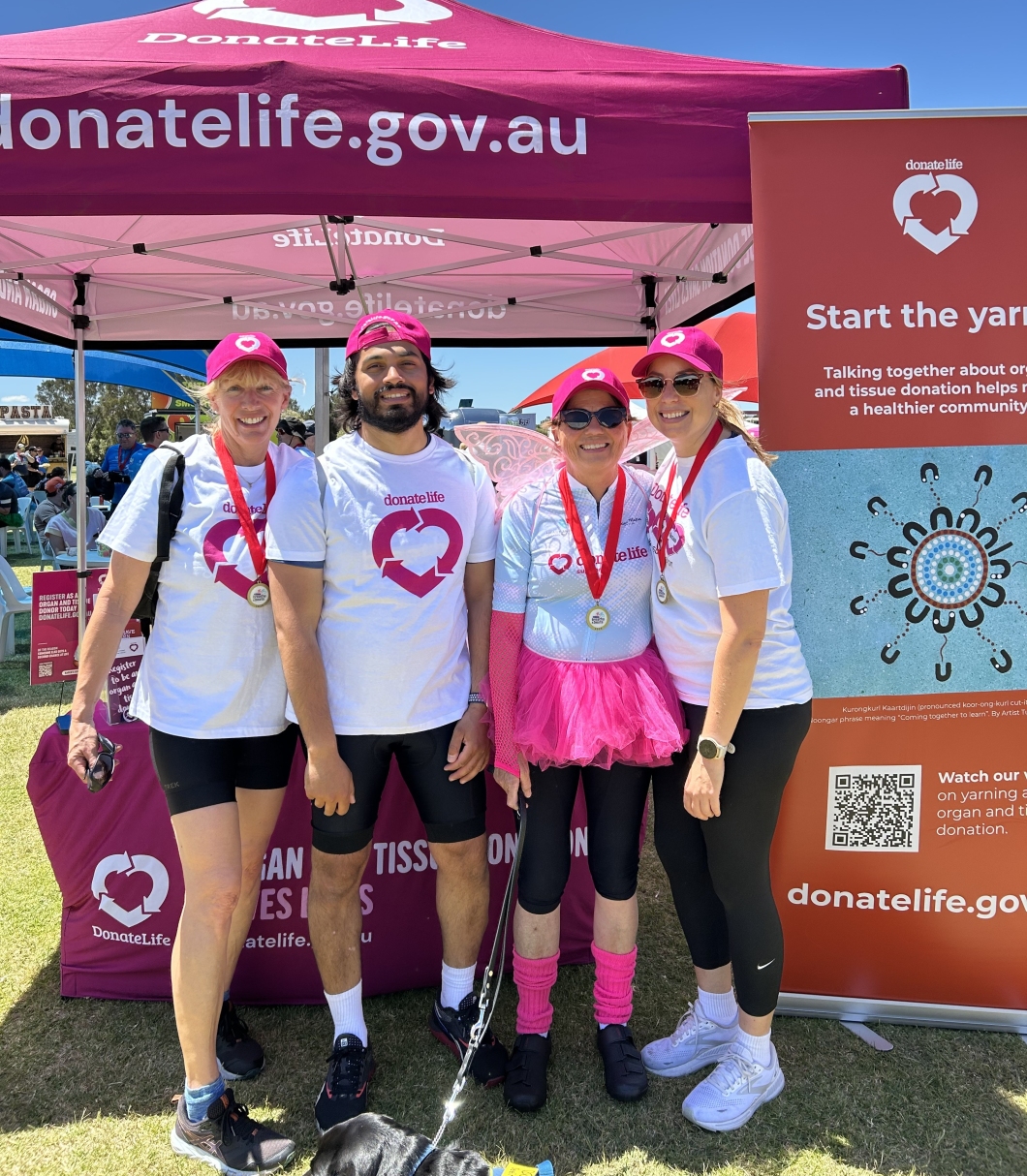4 people wearing DonateLife shirts and medals stand together in front of a bright pink DonateLife tent at an outdoor event. One person is dressed in a pink tutu and matching accessories. A DonateLife banner beside them reads ‘Start the yarn’ with information about organ and tissue donation. A dog on a leash is at their feet, and the scene is set on a sunny day with other event stalls visible in the background.