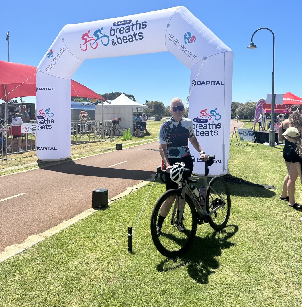A person stands with a bicycle under a large white inflatable event arch that reads ‘Breaths & beats.’ The scene is set on a sunny day at an outdoor cycling event, with tents, spectators, and a paved path visible in the background.