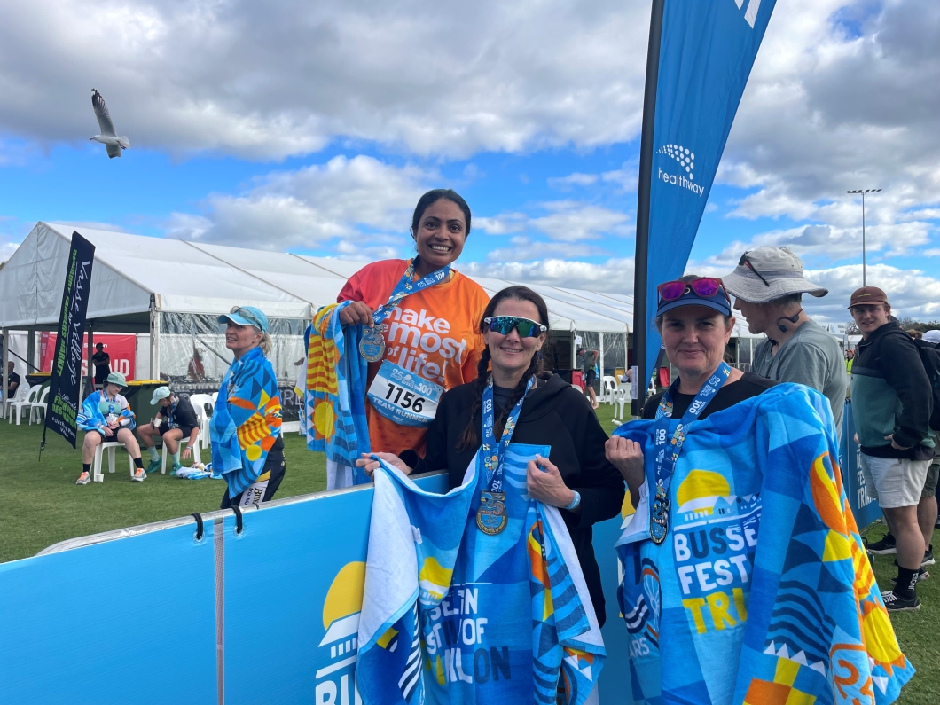 3 event participants stand outdoors at a finish area, wearing medals and holding colourful towels branded ‘Busselton Festival of Triathlon’. One person in an orange shirt with race bib number ‘1756’ displays a medal, while the others wear casual athletic clothing and sunglasses. White event tents, a blue Healthway banner, and other attendees are visible in the background under a cloudy sky.