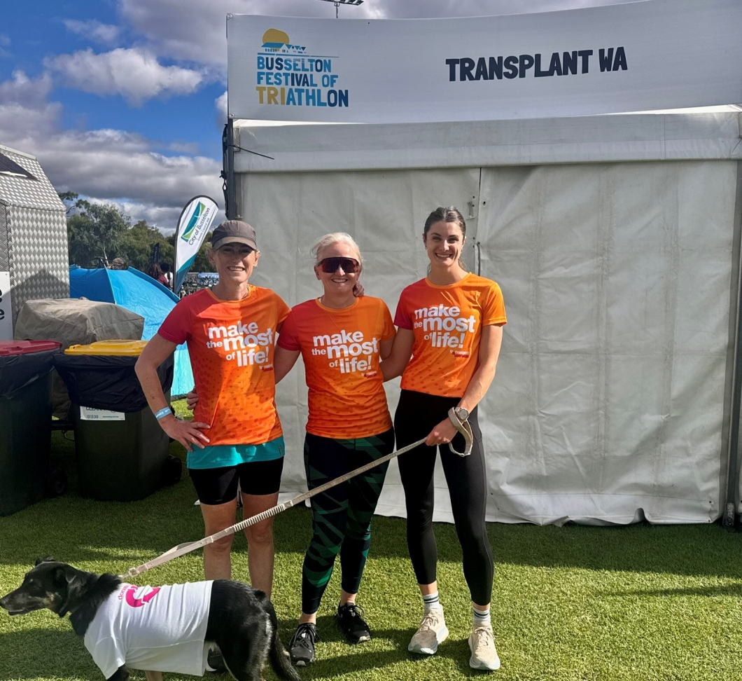3 participants stand on grass at the Busselton Festival of Triathlon in front of a white tent labeled ‘Transplant WA’. All 3 wear matching orange shirts with the text ‘Make the most of life!’ Two hold leashes attached to a black dog wearing a white vest with a DonateLife logo. Event tents, flags, and bins are visible in the background under a partly cloudy sky.