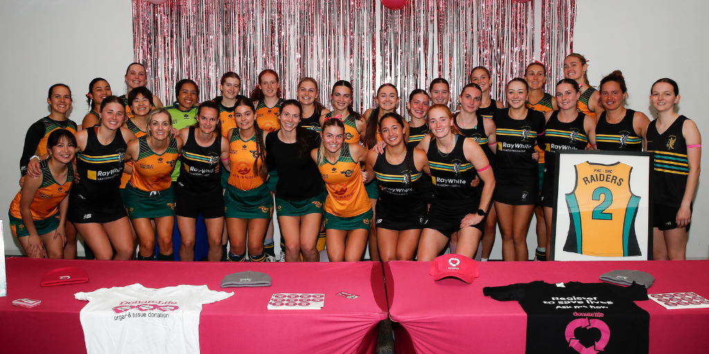 A large group of sports team members pose together indoors in front of a silver tinsel backdrop with red balloons. The group wears mixed team uniforms in green, gold, and black, featuring ‘Ray White’ branding and ‘Raiders’ logos. One framed jersey with the number 2 is displayed on the right. In the foreground, a table covered with a pink cloth holds DonateLife items, including caps, stickers, and shirts with text promoting organ and tissue donation, such as ‘Register to save lives’.