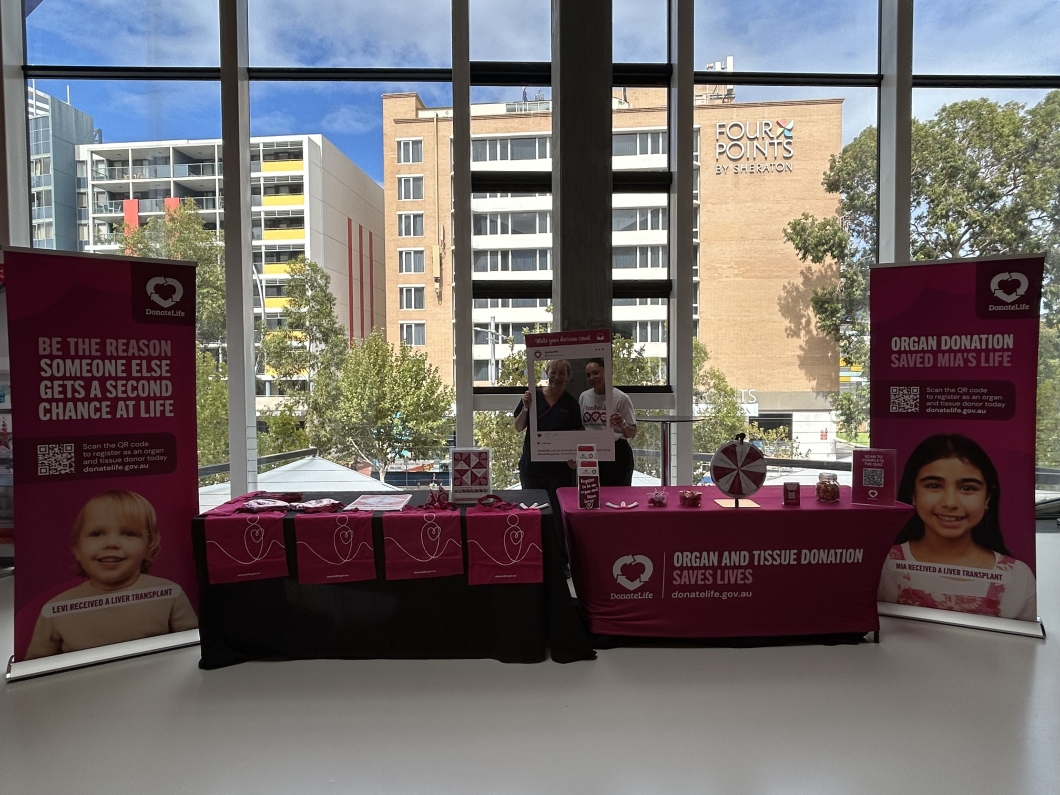 A DonateLife information display is set up indoors in front of large windows overlooking city buildings, including a visible ‘Four Points by Sheraton’ sign. Two people stand behind tables covered with magenta cloths displaying promotional materials, QR codes, and a spinning prize wheel. Tall magenta banners on either side read messages such as, ‘Be the reason someone else gets a second chance at life’ and ‘Organ and tissue donation saves lives’, alongside images of transplant recipients.