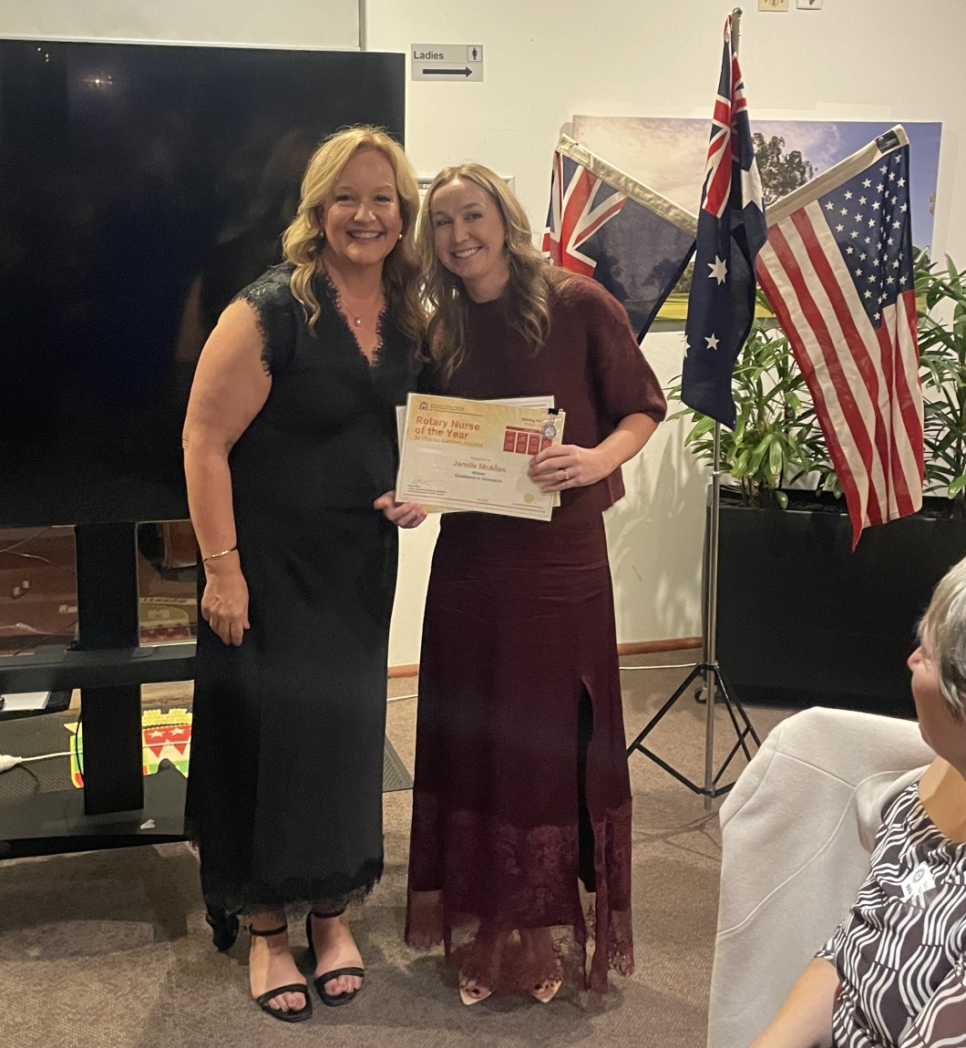 2 people stand indoors at an awards presentation, posing together while holding a framed certificate titled ‘Rotary Nurse of the Year’. The setting includes a large TV screen and Australian and U.S. flags displayed on stands behind them. One person wears a black dress, while the other wears a burgundy dress and holds the certificate.