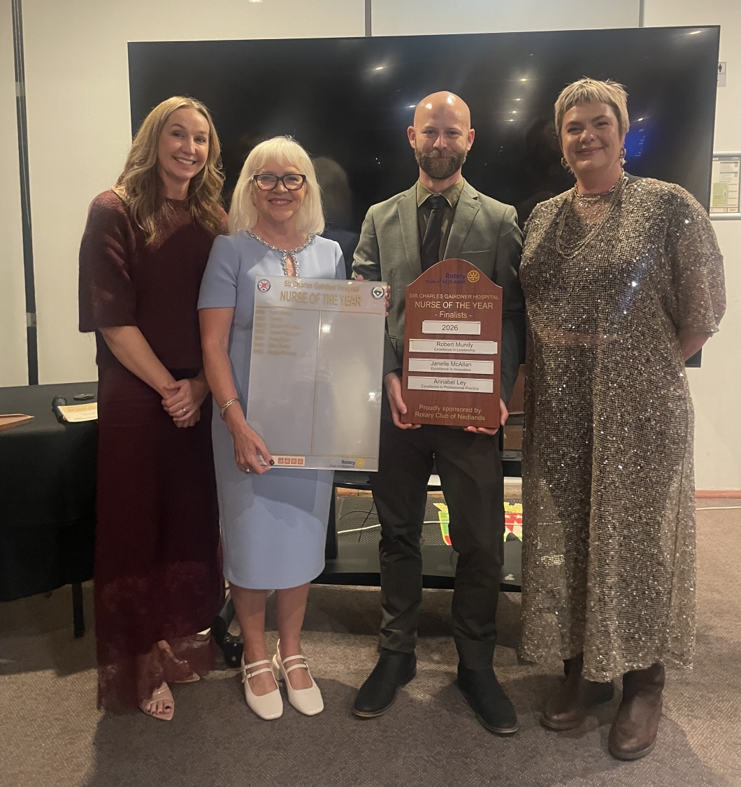4 adults stand indoors in front of a large screen, posing together at an awards presentation. Two people in the centre hold a large ‘Nurse of the Year’ board and a wooden plaque listing finalists and the year 2026, sponsored by Rotary Club of Nedlands. The group is dressed in formal attire, including dresses and a suit.