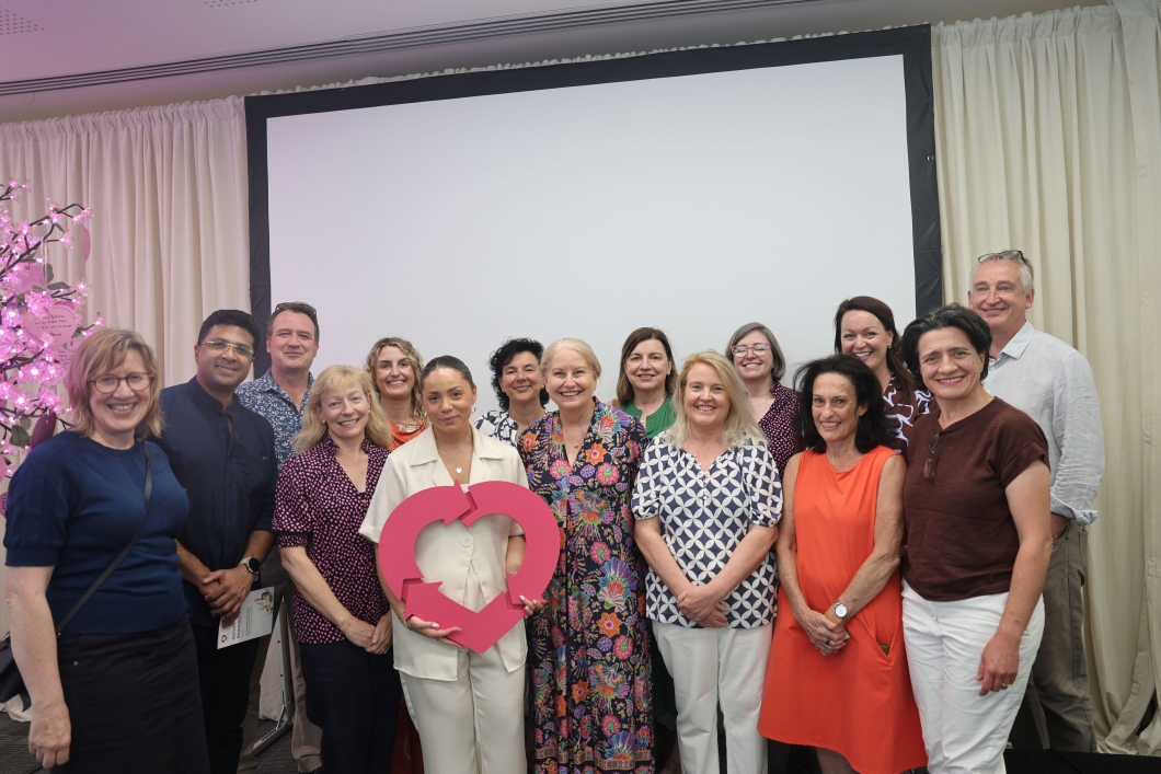 A group of people stand together indoors in front of a large blank projection screen. One person at the front holds a large pink heart-shaped DonateLife symbol. Soft lighting and decorative elements, including illuminated branches on the left, frame the scene.