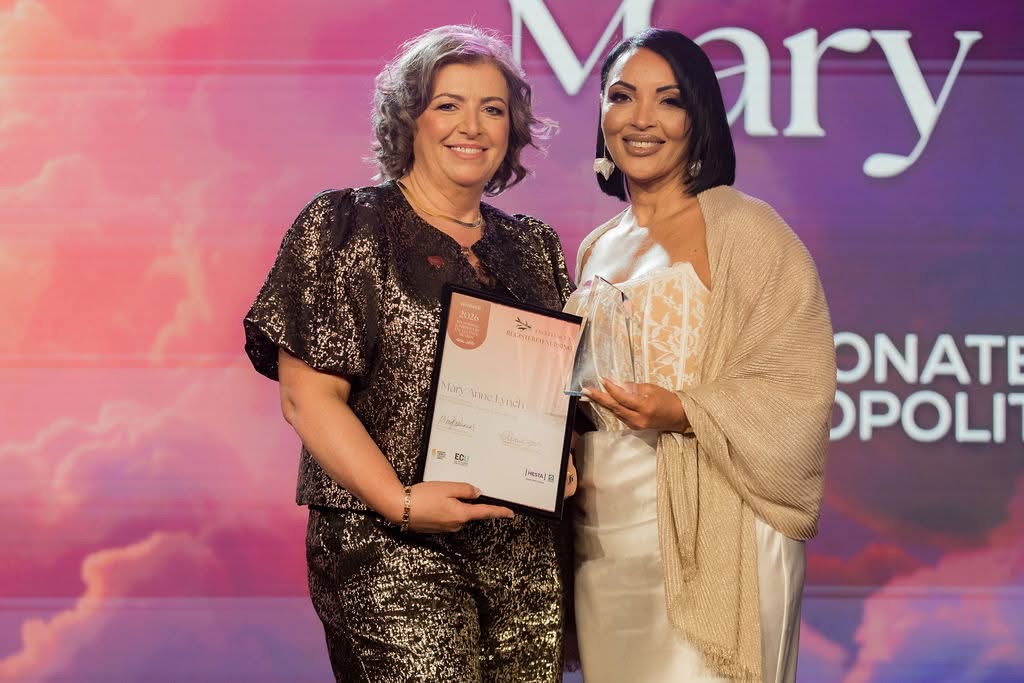 2 people stand on a stage in front of a purple and pink event backdrop, holding an award and a framed certificate. One person wears a dark, sparkly outfit, while the other wears a light satin dress with a shawl and holds a glass trophy. The certificate includes visible text, and the backdrop features large lettering, including the word ‘Mary’.