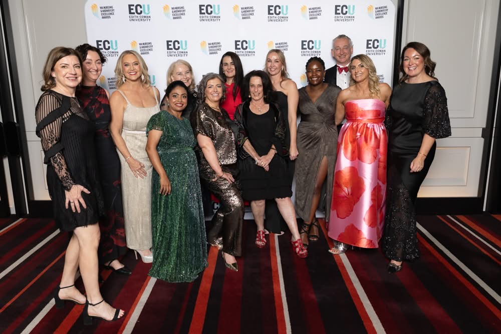 : A group of 13 adults pose together on a striped carpet in front of a step-and-repeat backdrop displaying ‘ECU Edith Cowan University’ and ‘WA Nursing Excellence Awards’ logos. The group is dressed in formal attire, including dresses, suits, and evening wear in black, red, green, and metallic tones. One person wears a bright pink-and-red patterned gown, while another wears a green embellished dress. The backdrop and lighting indicate an indoor awards event setting.