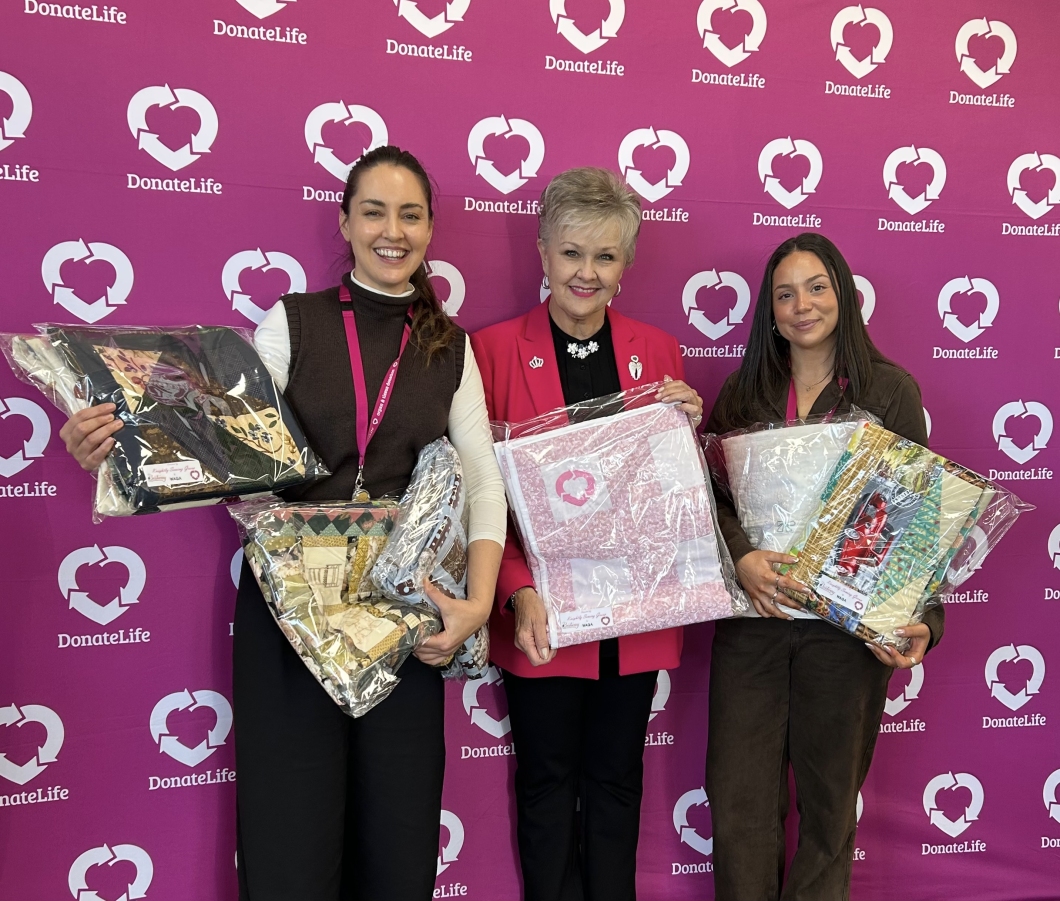 3 people stand in front of a magenta DonateLife backdrop, holding handmade quilted items wrapped in plastic. The display showcases community-crafted quilts created to support and raise awareness of organ and tissue donation during a DonateLife event.
