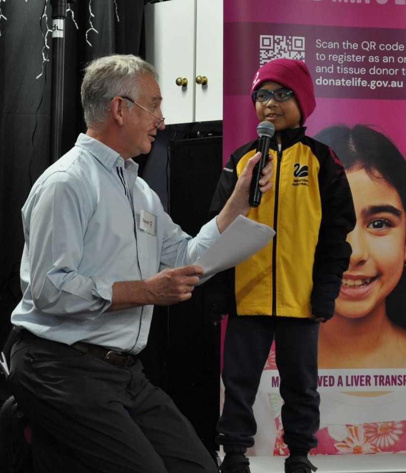 A child wearing a magenta beanie speaks into a microphone on stage beside a DonateLife banner, while an event host kneels nearby holding notes during a community awareness event.