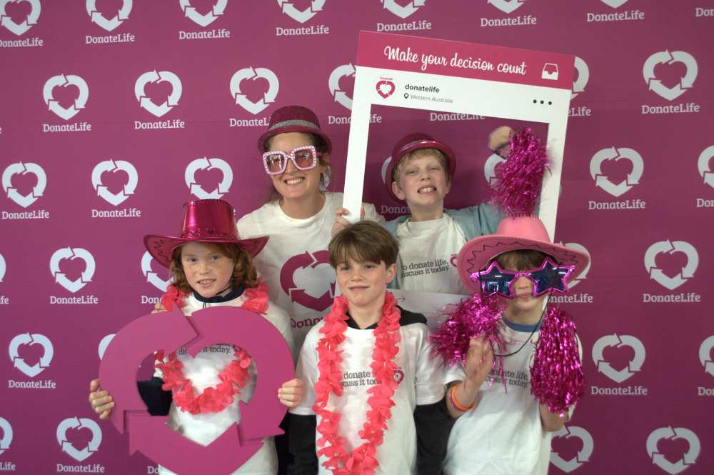 An adult and 4 children pose in front of a magenta DonateLife backdrop, wearing colourful costume accessories and holding a large magenta DonateLife symbol and a photo-frame prop during a community event.