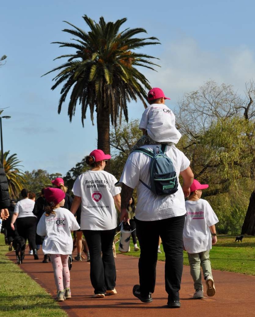 Participants wearing white DonateLife shirts and magenta caps walk along a shared path lined with trees during a community Lap for Life event promoting organ and tissue donation.