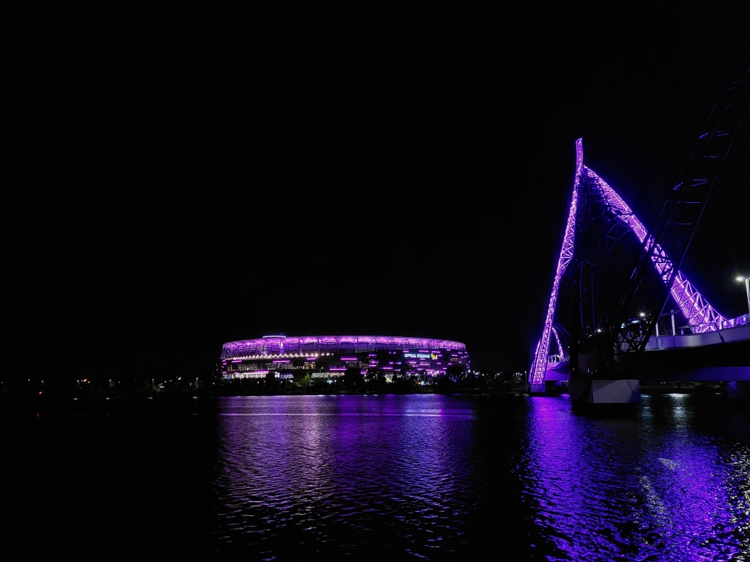 Optus Stadium and the Matagarup Bridge are illuminated in magenta at night, with their reflections visible on the Swan River.