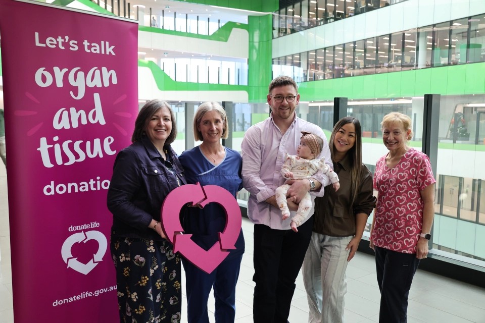 A group of healthcare staff and community members stand beside a magenta DonateLife banner in a hospital atrium, holding a large magenta DonateLife symbol during an organ and tissue donation awareness event.