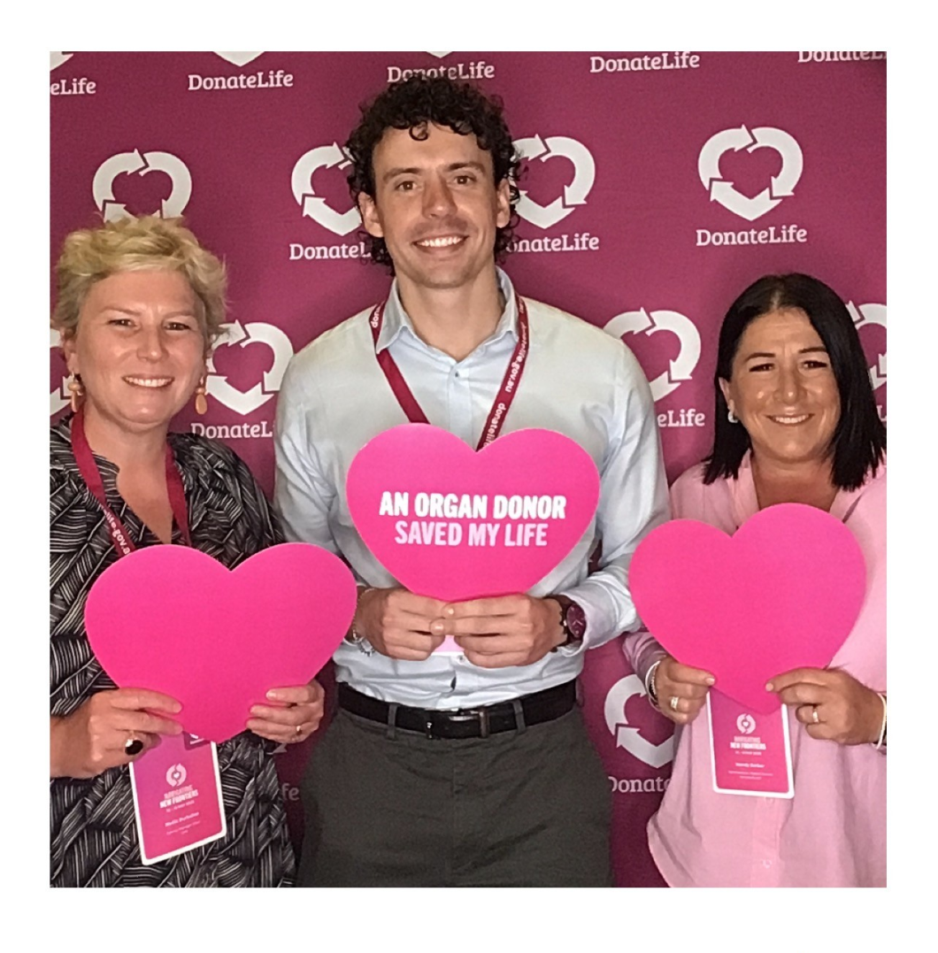 Three people hold large pink hearts, the middle one with the message, ‘An organ donor saved my life’, against a DonateLife backdrop.
