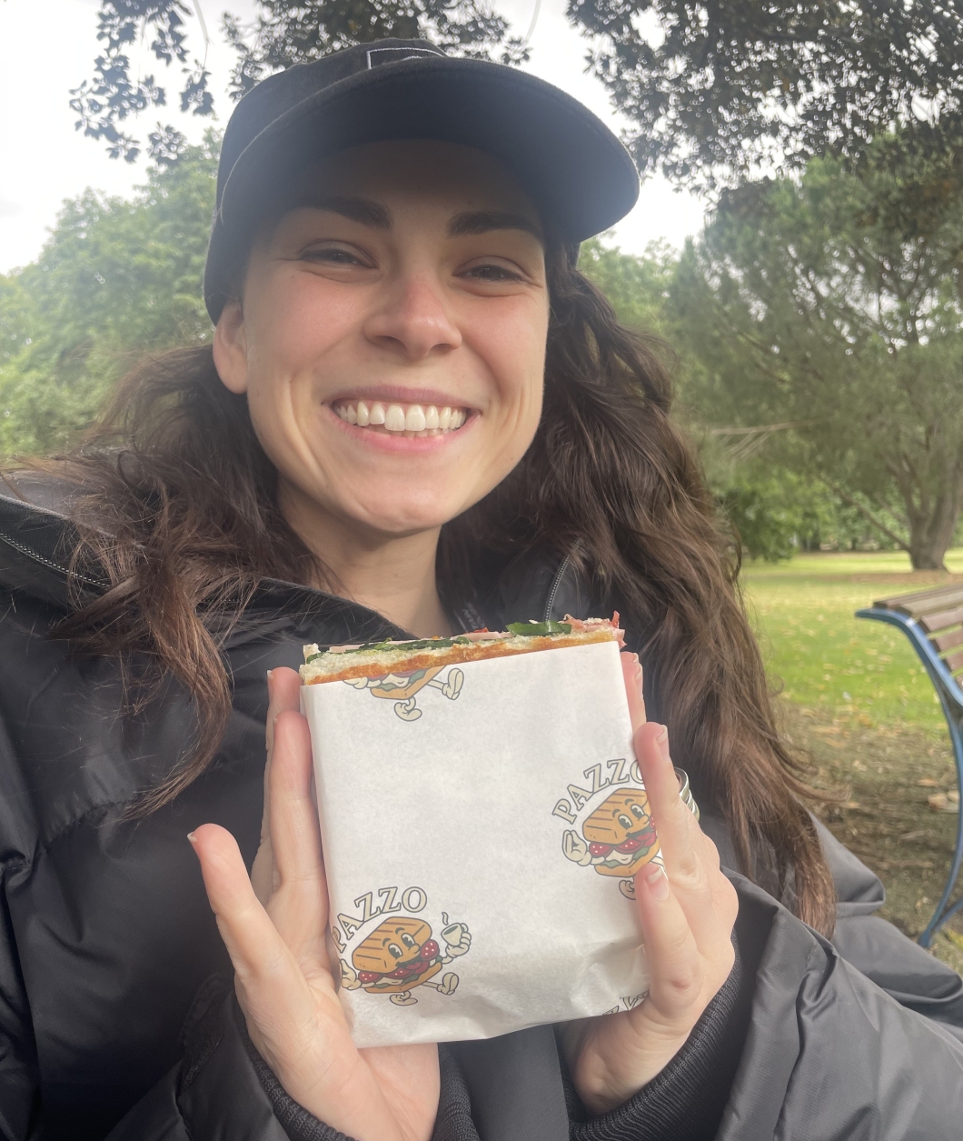 A person sits outdoors on a park bench, holding a wrapped sandwich with ‘PAZZO’ branding on the paper. They are wearing a dark puffer jacket and a cap, with trees and greenery visible in the background.