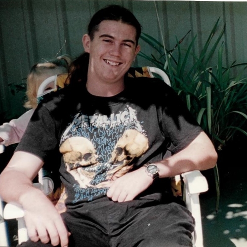 Doug is in a black shirt with skulls, sitting in a lawn chair smiling at the camera. Green fence and shrubbery in the background. A small girl is half hidden behind Doug's chair.