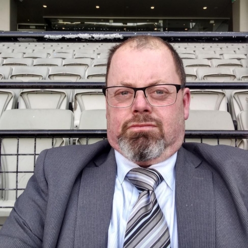 Tim in a suit and tie sitting in a sport stadium chair looking at the camera