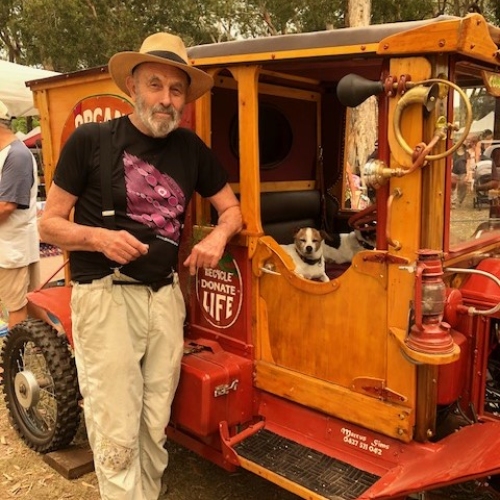 Man standing next to an old orange automobile which has two little dogs sitting in the front seat. 