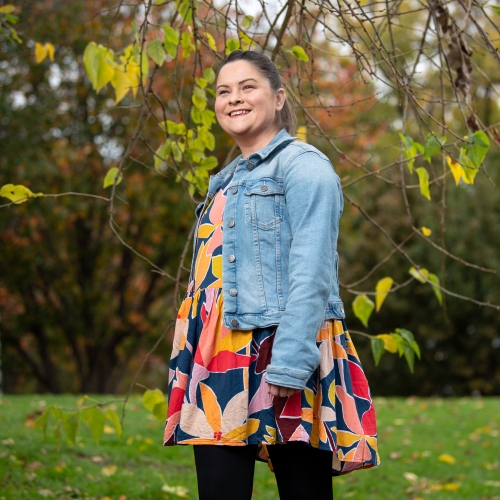 Woman in a bright patterned dress and a denim jacket. She is standing outside on a green lawn surrounded by green leaves.