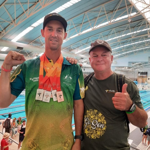 Two men at an indoor swimming pool. One has a couple of medals around his neck.