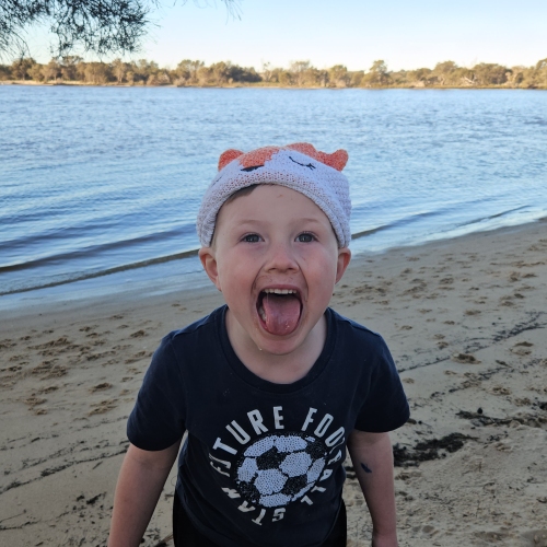Little boy in a black shirt and white beanie, sticking his tongue out in a playful manner towards the camera. He is standing outside on a sandy bank with a lake in the background.
