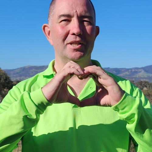 Man in a bright green shirt holding his hands in the shape of a heart. He is standing outside and you can see a large expanse of blue sky and mountains in the background.