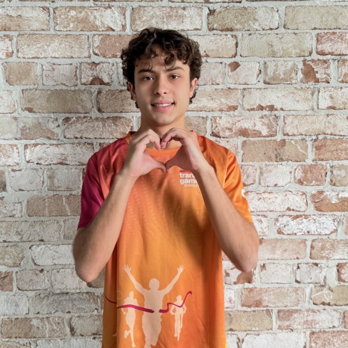 Young man in an orange shirt, standing in front of a brick wall, holding his hands in the shape of a heart.