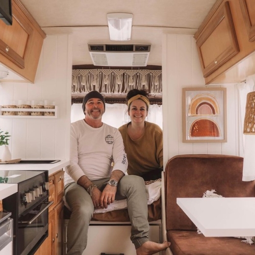 a man and a woman sitting inside a travel van smiling at the camera. The inside of the van is clean, white and has wooden cabinets lining the top of the van.